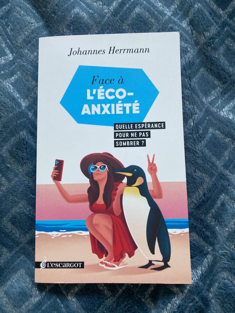 Photo de la couverture du livre de Johannes Herrmann intitulé "Face à l'éco anxiété". On y voit un dessin d'une femme en robe rouge, capeline rouge et lunettes de soleil qui est accroupie sur la plage à côté d'un côté d'un manchot. Elle prend un selfie avec son téléphone portable en faisant le signe V de victoire derrière la tête du manchot.