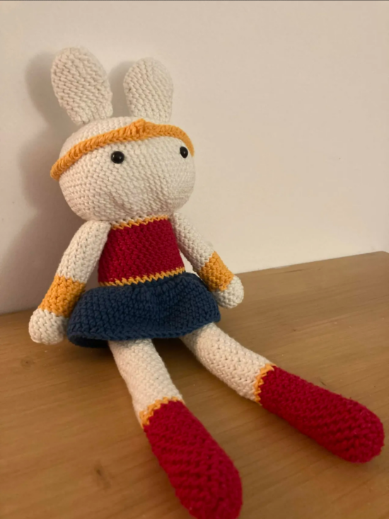 Photo d'un amigurumi de lapin avec un costume de Wonder Woman. La photo est prise de trois quarts, le lapin regarde sur la droite. Il est adossé à un mur blanc et assis sur un bureau en bois clair.