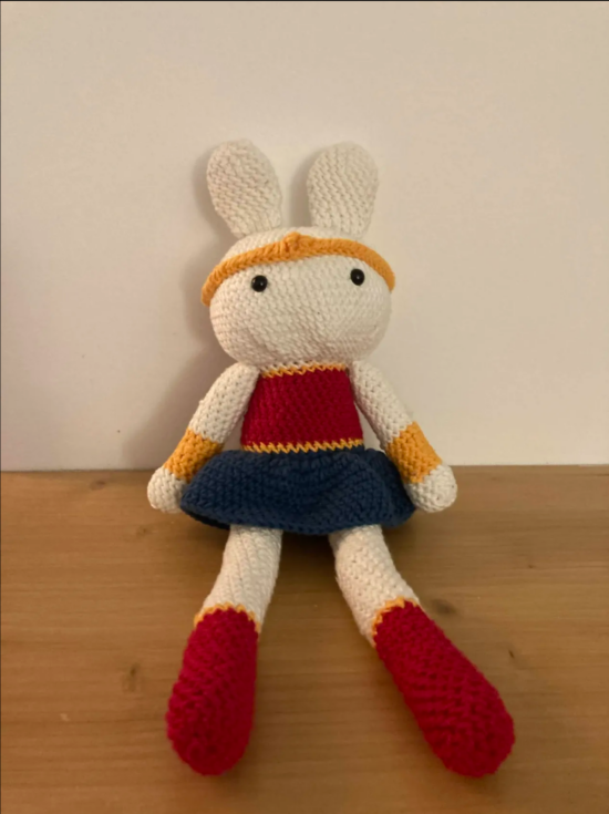 Photo d'un amigurumi au crochet d'un lapin avec un costume de wonderwoman. La photo est prise de face, le lapin est adossé à un mur blanc et assis sur un bureau en bois clair.