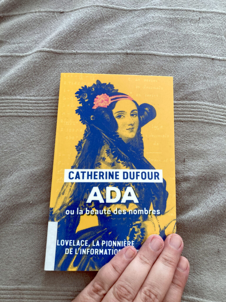 Photo du livre intitulé Ada ou la beauté des nombres de Catherine Dufour, en édition poche. Le livre est jaune avec un portrait d'Ada Lovelace en bleu. Le livre est posé sur un tissu gris texturé, ma main gauche est posée sur le coin en bas à droite du livre