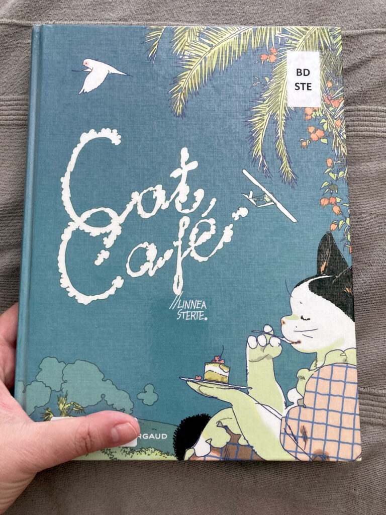 Photo de la couverture de la BD Cat Café. On y voit un chat blanc et noir vêtu d'une robe rose à carreaux bleus et des froufrous assis et en train de déguster une pâtisserie. Le chat est assis dehors et dans le ciel bleu un hydravion vole.