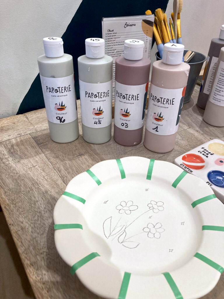 Photo de la céramique que j'ai choisie, une petite assiette qui ressemble à une fleur. J'ai dessiné des fleurs au centre de l'assiette et j'ai également positionné des petits scotchs pour peindre proprement. Sur la table, on voit également des tubes de couleur, des pinceaux, un bout de palette et on distingue un morceau de la carte des boissons.