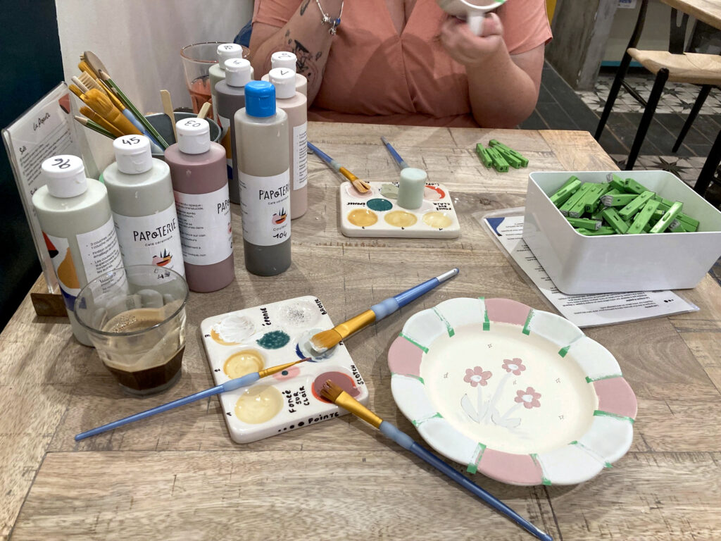 Photo de ma production en cours de décoration. Sur la table, outre l'assiette que je décore, on voit les pots de couleur, un verre de café, un verre de limonade, un pot avec des tampons en forme de lettres, des palettes et des pinceaux. En arrière plan, Paupoune est en train de peindre son mug.