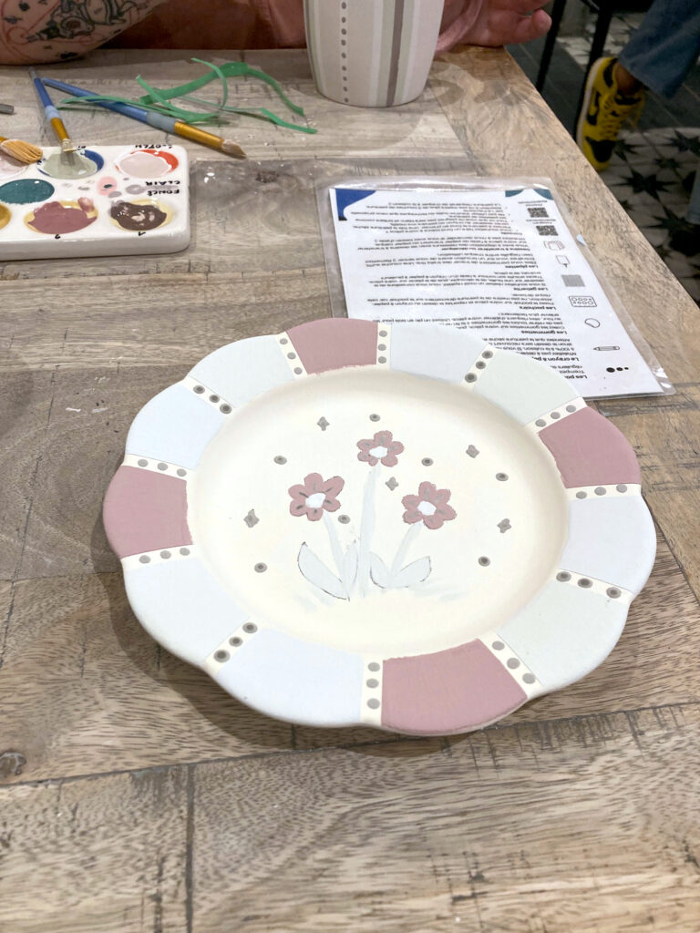 Photo de ma production posée sur une table. Il s'agit d'une assiette avec un bord qui rappelle une fleur. J'ai coloré les pétales dans les tons pourpres, bleus et verts. Entre chaque couleur, des pois qui après cuisson devraient être en paillettes dorées. Au centre de l'assiette, trois fleurs de type marguerites, entourées de paillettes et pois. Les couleurs sont pastel car la pièce n'a pas encore été cuite.