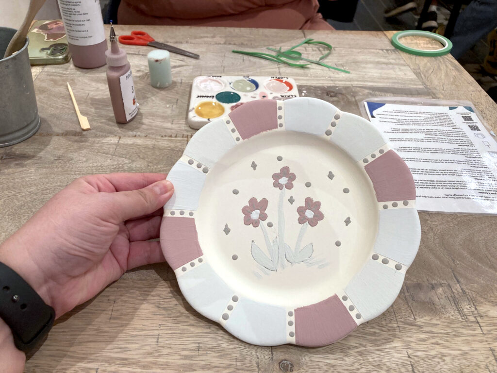 Photo de ma production que je tiens dans ma main. Il s'agit d'une assiette avec un bord qui rappelle une fleur. J'ai coloré les pétales dans les tons pourpres, bleus et verts. Entre chaque couleur, des pois qui après cuisson devraient être en paillettes dorées. Au centre de l'assiette, trois fleurs de type marguerites, entourées de paillettes et pois. Les couleurs sont pastel car la pièce n'a pas encore été cuite.