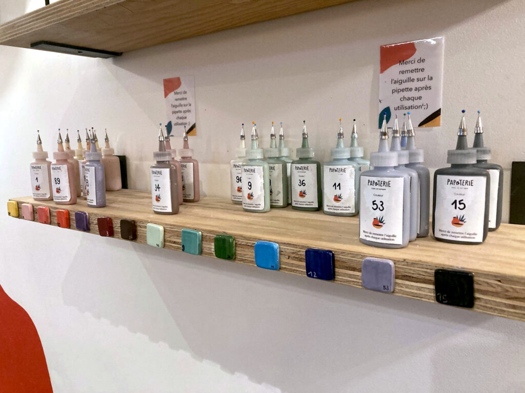 Photo d'une étagère de la Papoterie avec des fioles alignées et rangées par couleur. Il y a aussi des petits morceaux de céramique qui montrent la couleur finale après cuisson des différentes teintes proposées. Il y a sur le mur de petits écriteaux qui précisent "Merci de remettre l'aiguille sur la pipette après chaque utilisation".