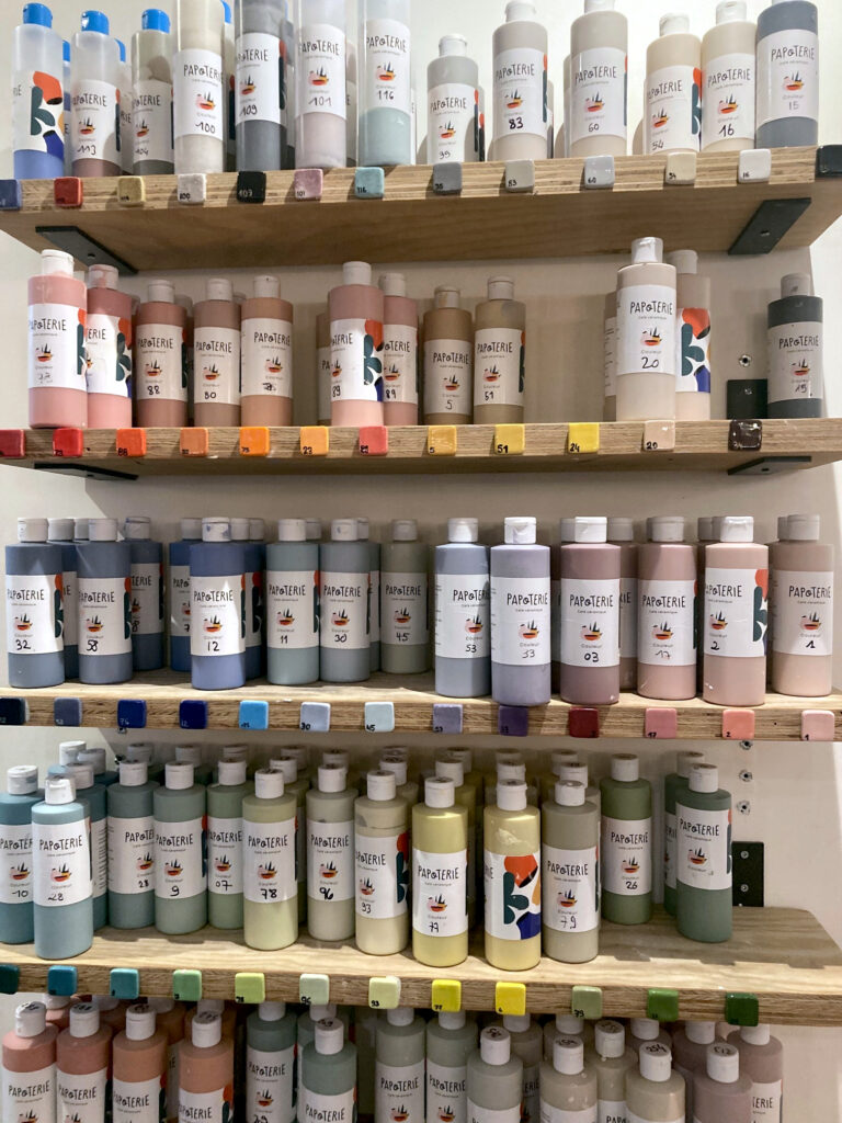 Photo d'un pan de mur de la Papoterie avec des étagères sur lesquelles sont disposées des contenants avec de la peinture à céramique. Les contenants sont rangés par couleur et le rendu final de la couleur après cuisson est positionné sur un petit carreau de céramique fixé sur l'étagère, en dessous des contenants.