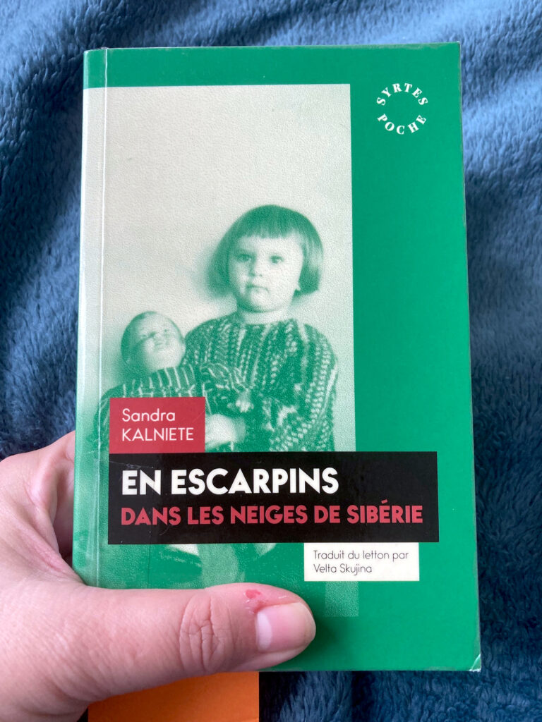 Photo de la couverture du livre de Sandra Kalniete intitulé "En escarpins dans les neiges de Sibérie". La couverture est de couleur verte avec une photo de l'autrice enfant qui tient un poupon dans ses bras.